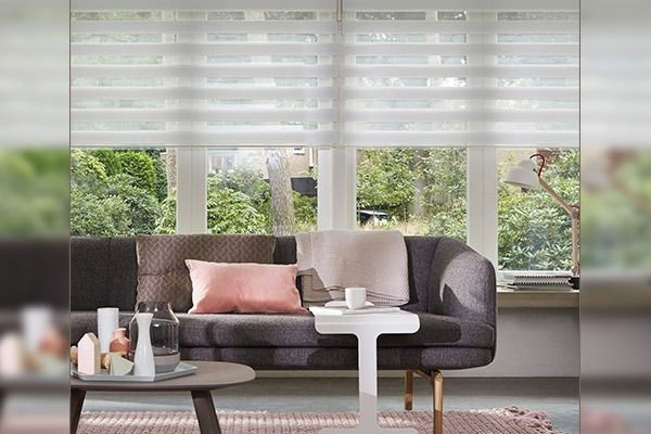 Salón con sofá gris oscuro, cojines rosa claro y persianas blancas superpuestas.