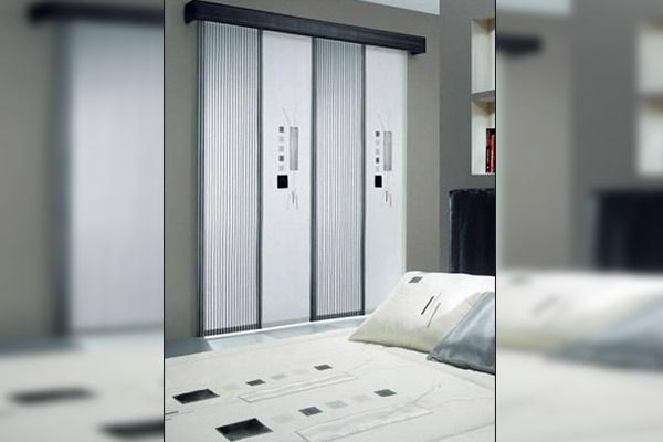 Puertas correderas de ascensor en una habitación de color claro, parcialmente abiertas. Una cama con almohadas en primer plano.