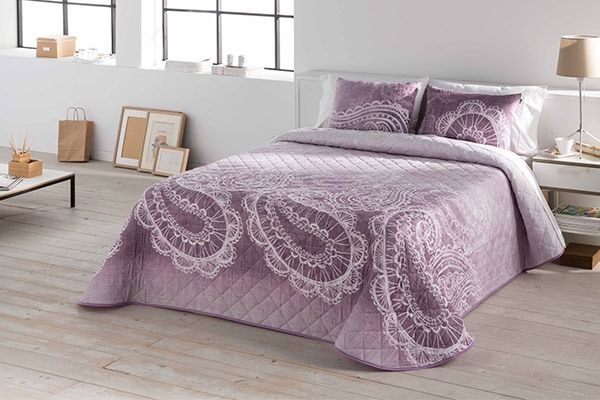 Colcha y almohadas color lavanda con estampado paisley blanco sobre un fondo blanco.