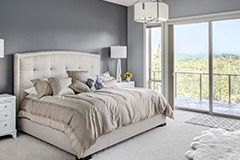 Dormitorio con pared gris, cama grande con ropa de cama beige y puerta corredera de cristal con vistas a los árboles.