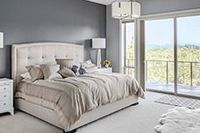 Dormitorio con pared gris, cama grande con ropa de cama beige y puerta corredera de cristal con vistas a los árboles.