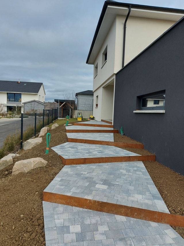 Allée de jardin pavée longeant une maison moderne, avec des dalles de pierre grise surélevées.