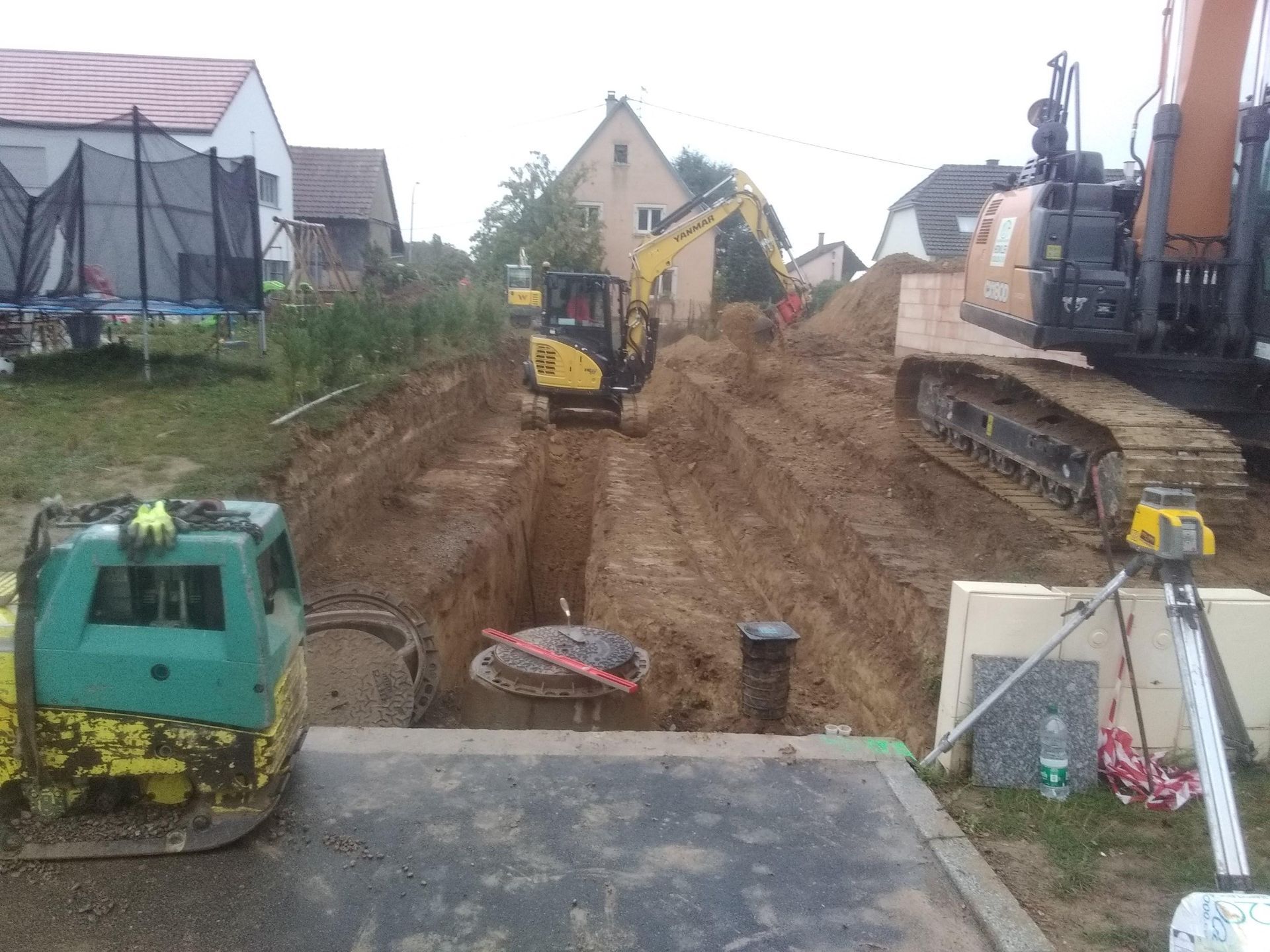 Chantier de construction avec des excavatrices creusant une tranchée entre des maisons.