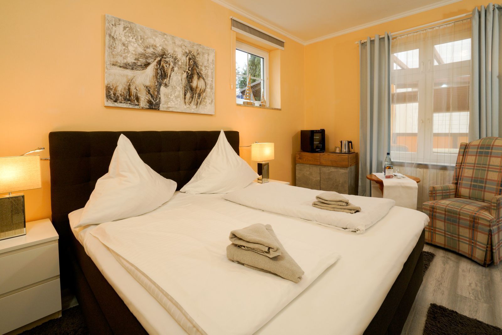 Schlafzimmer mit einem großen Bett, zwei Nachttischen, einem Stuhl und Kunstwerken an der Wand; hellorange Wände.