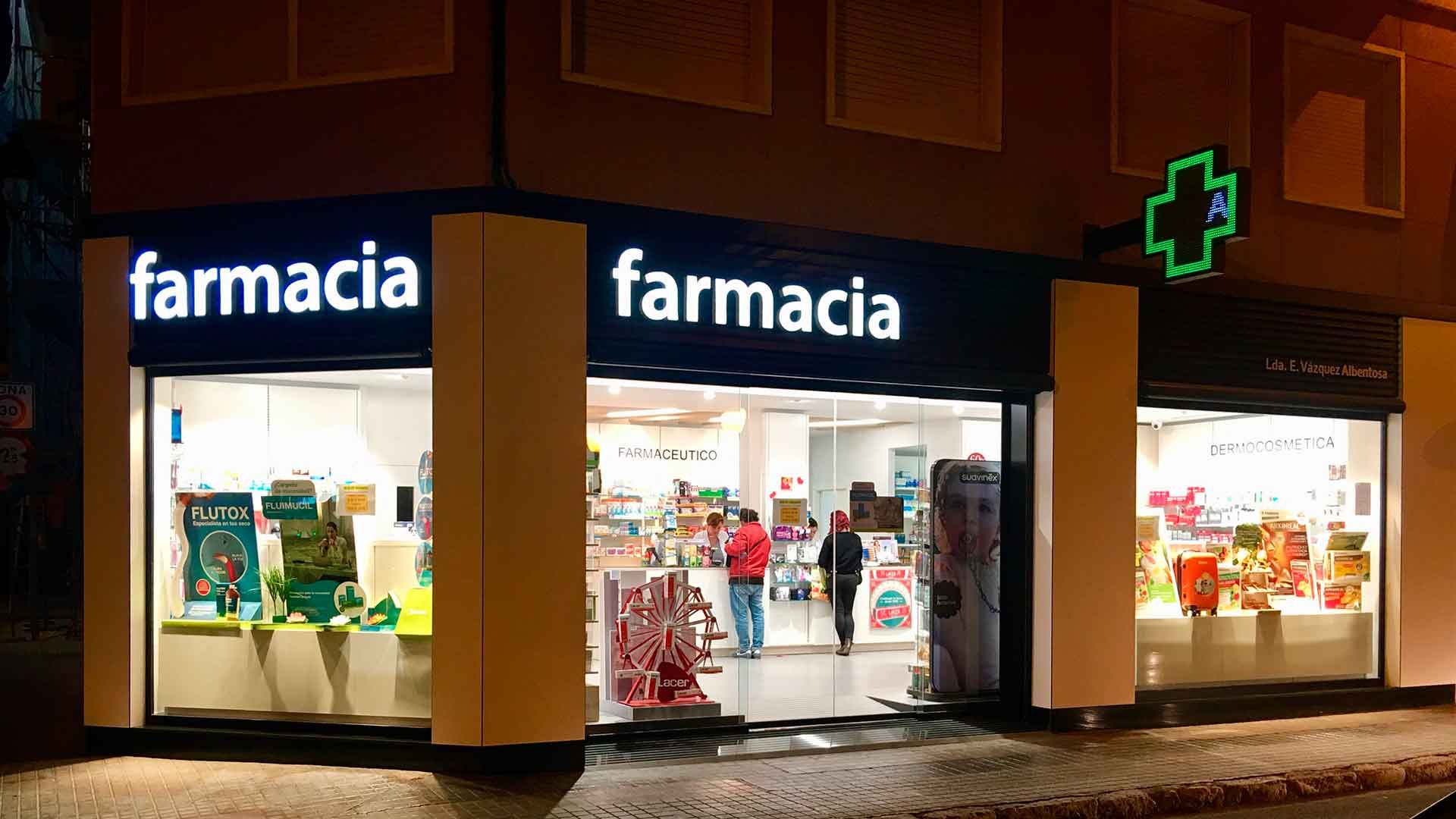 Una farmacia está iluminada por la noche con gente parada frente a ella.