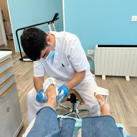 En una clínica, un profesional sanitario con bata y mascarilla aplica una sustancia blanca en los pies de un paciente.