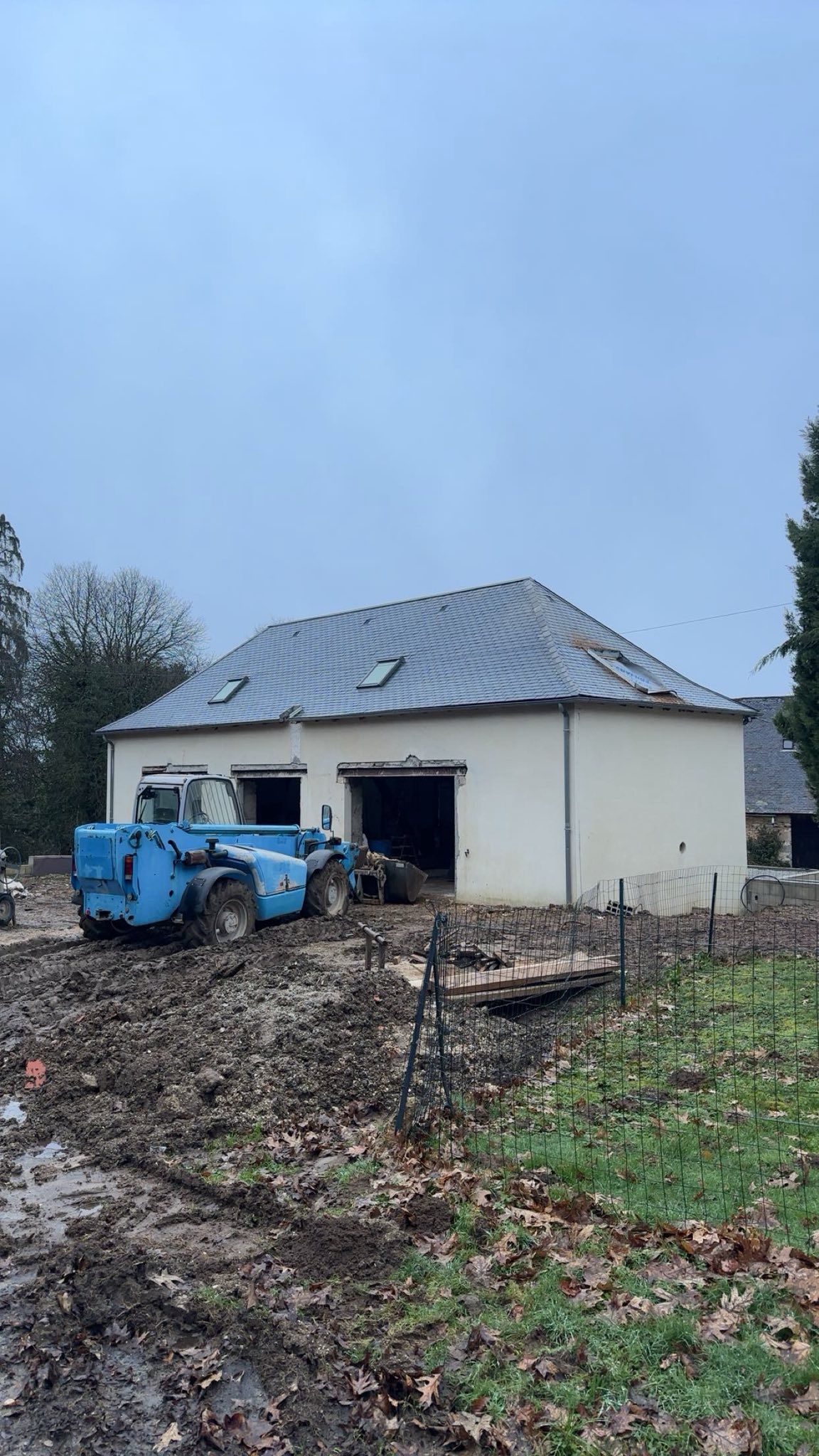 Engin de terrassement bleu devant une maison blanche à la cour boueuse.