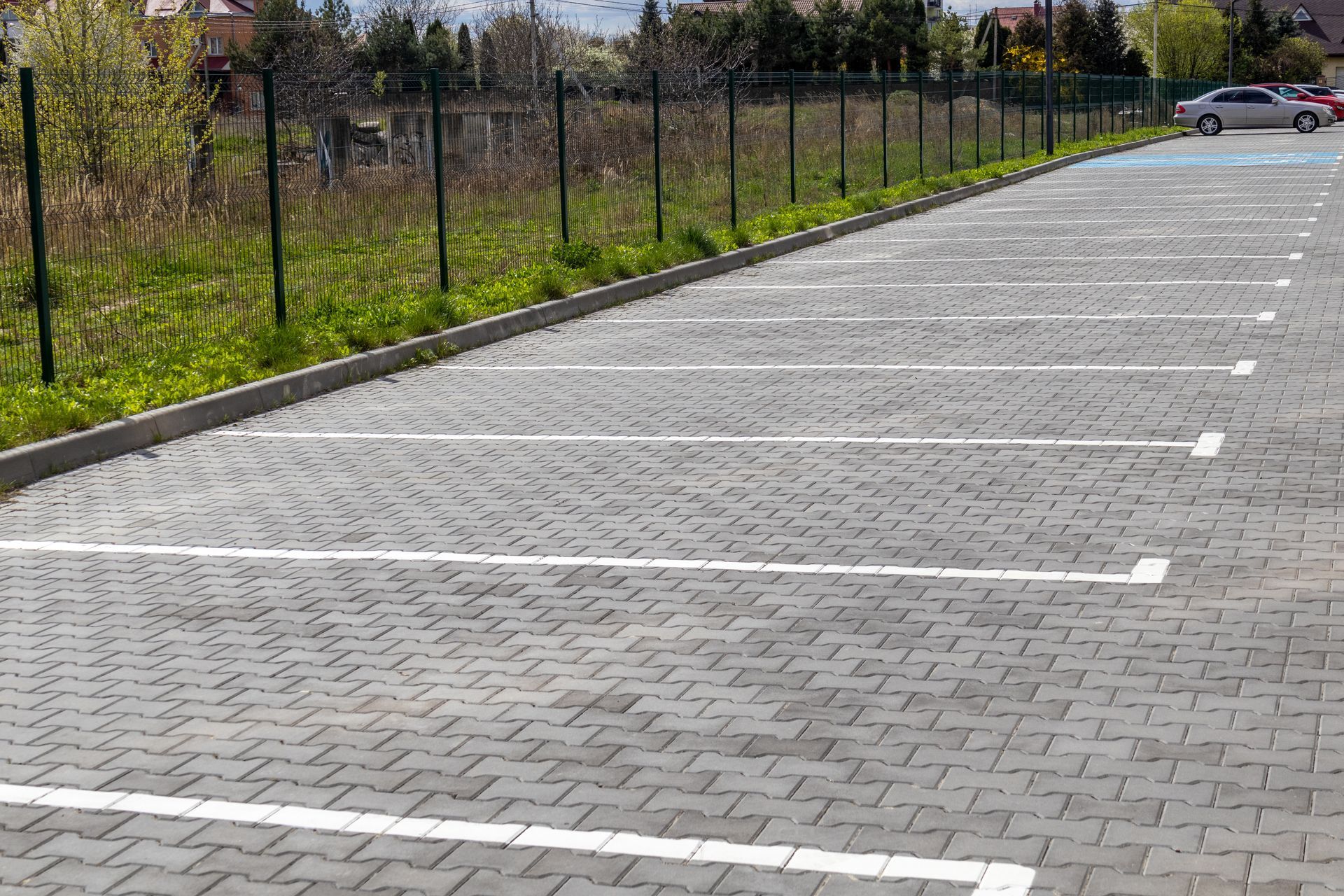 Parking goudronné vide, bordé d'une clôture, avec des lignes blanches et quelques voitures au loin.