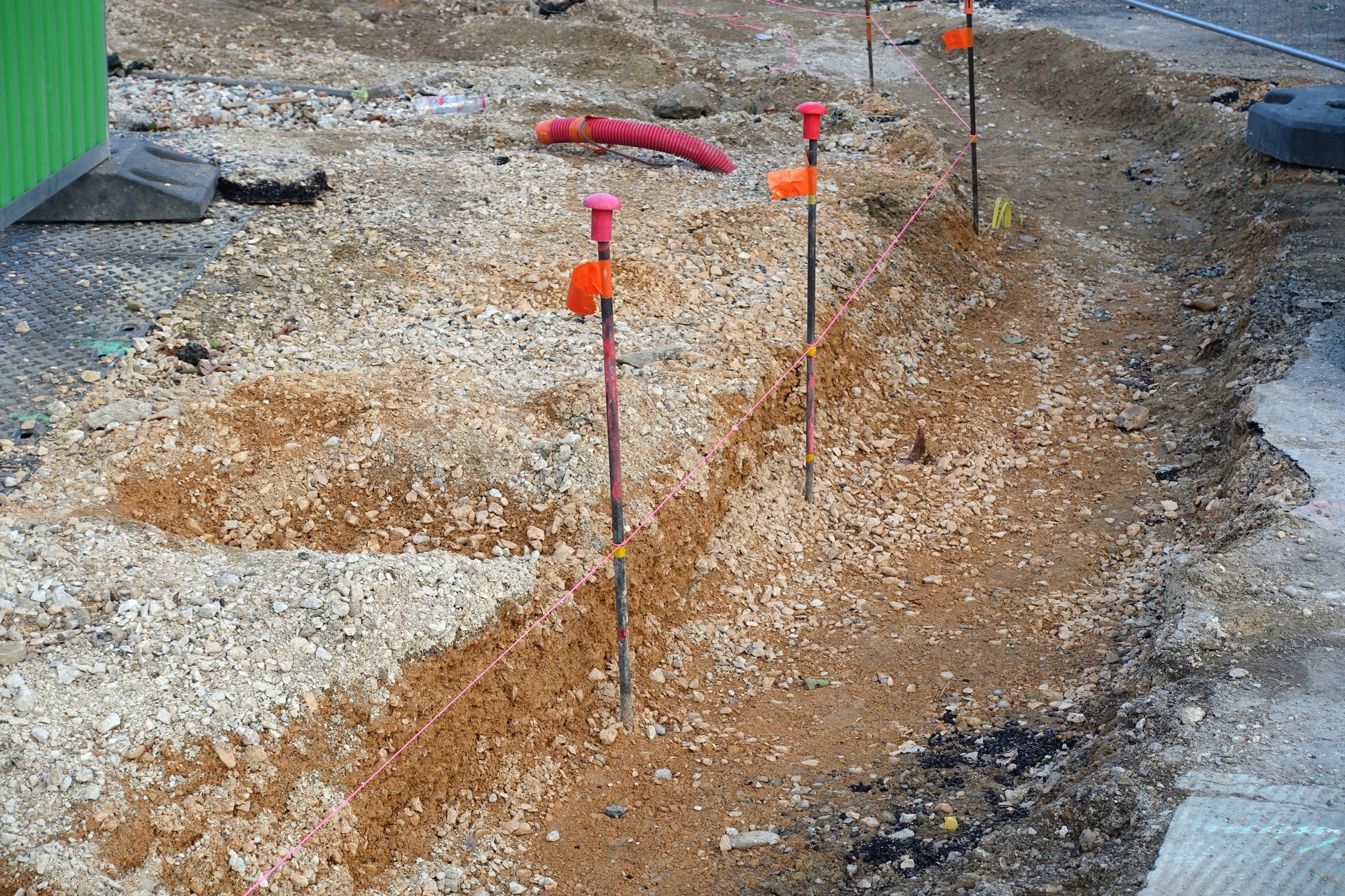 Tranchée technique avec des piquets de géomètre orange et du gravier à côté d'un chantier routier.