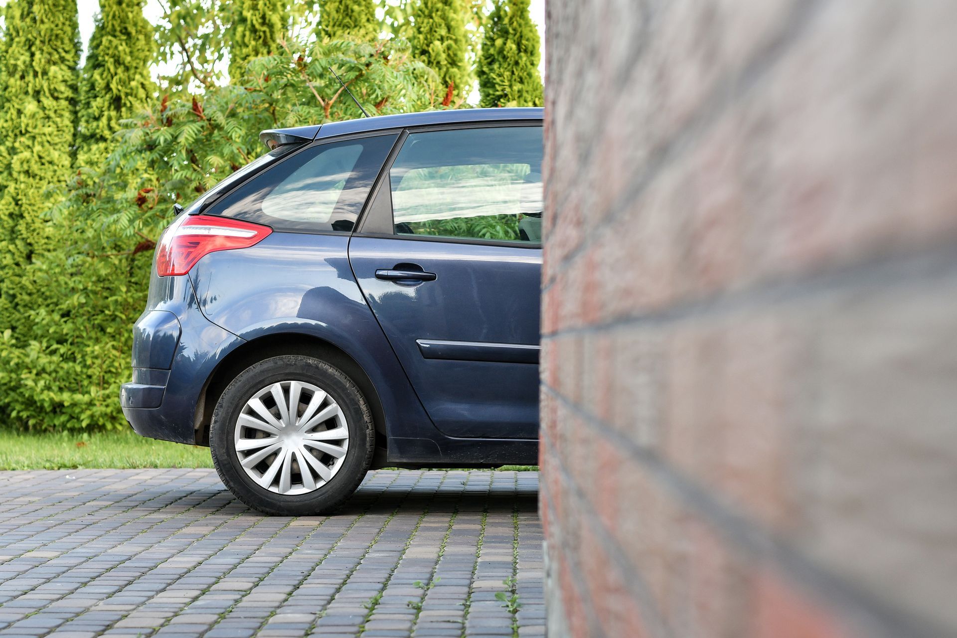 Voiture bleue garée sur une allée pavée, à côté d'un mur de briques et de verdure.