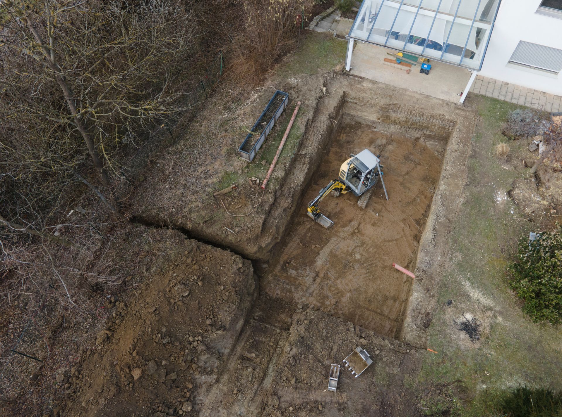 Vue aérienne d'une excavation dans un jardin à côté d'une maison, avec une minipelle et des tas de terre.
