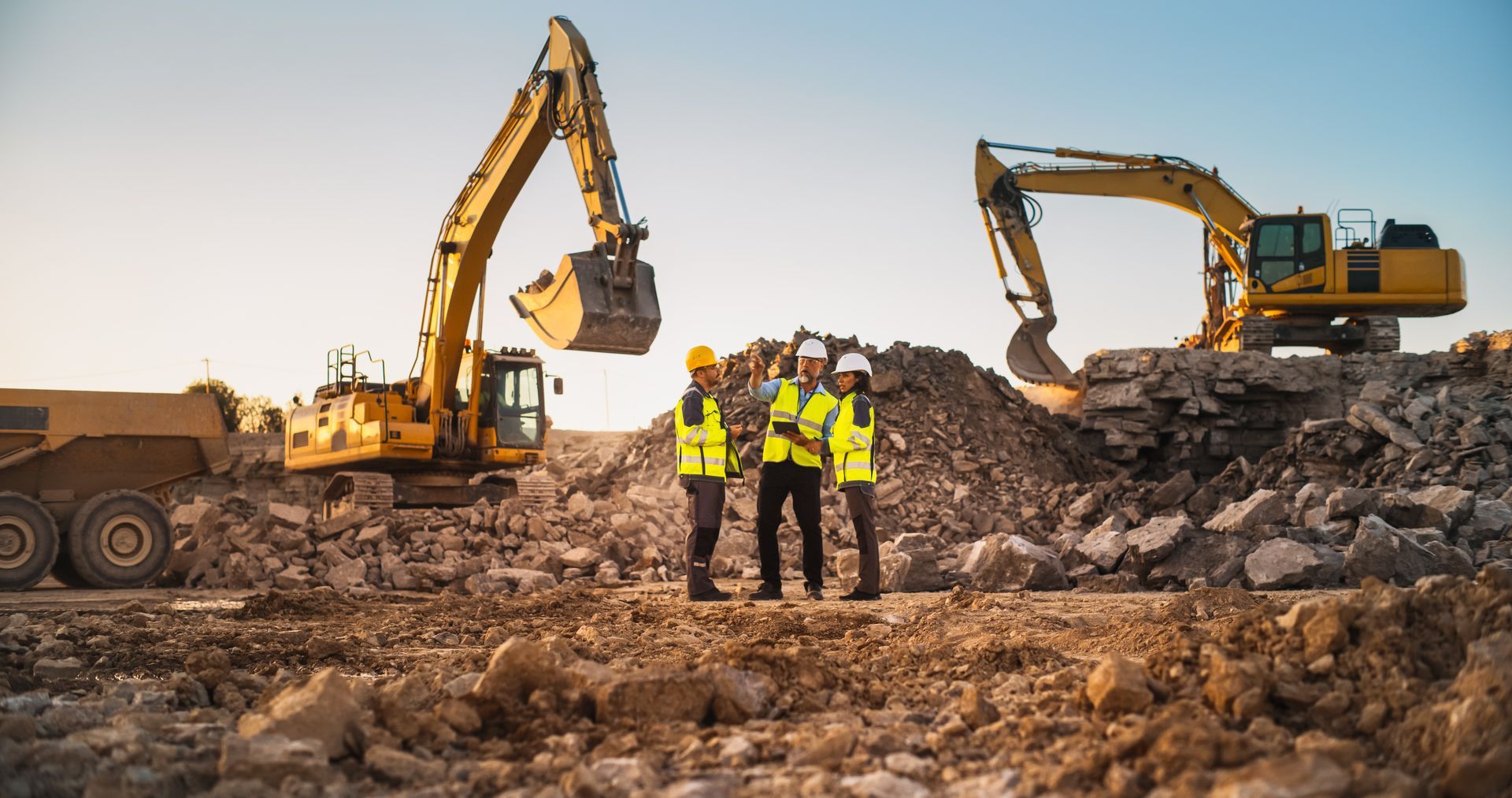 Des ouvriers du bâtiment en gilets jaunes se tiennent entre deux excavatrices sur un chantier rocailleux.