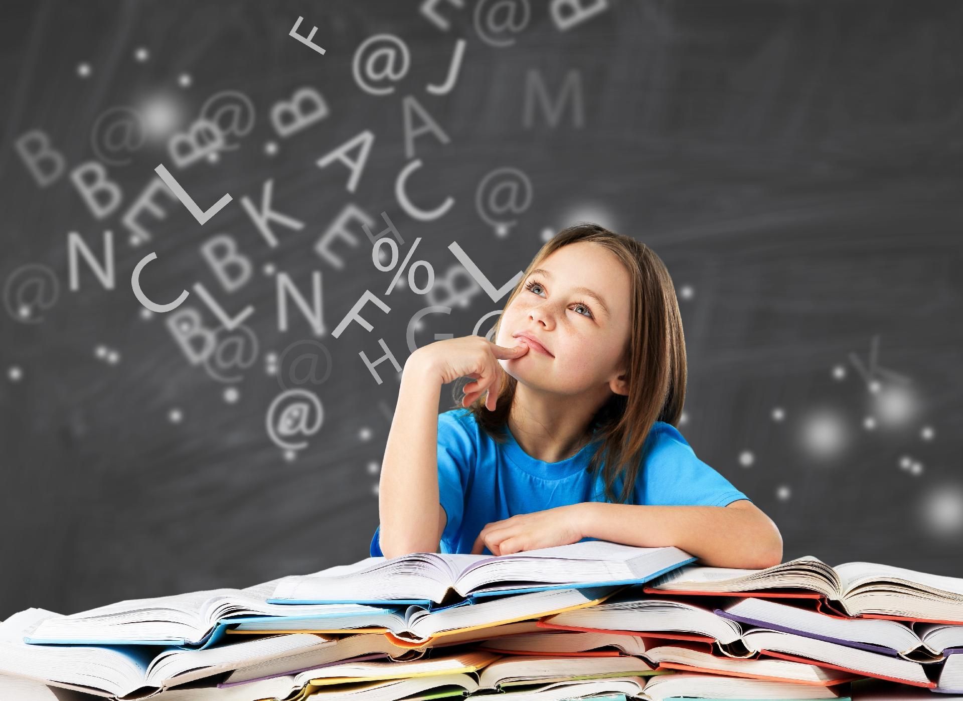 Mädchen sitzt am Tisch und blickt nachdenklich vor eine Tafel mit Buchstaben.