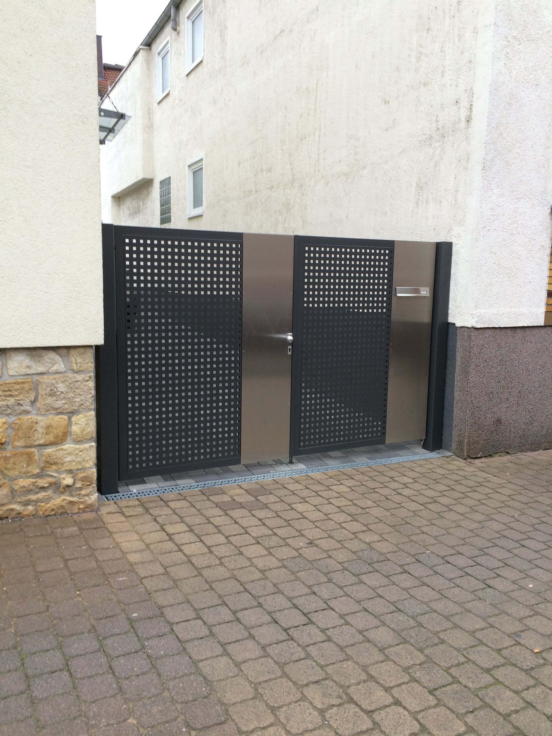 Schwarzes, perforiertes Metalltor an einem Wohnhauseingang neben Stein- und weißen Mauern auf einem gepflasterten Weg