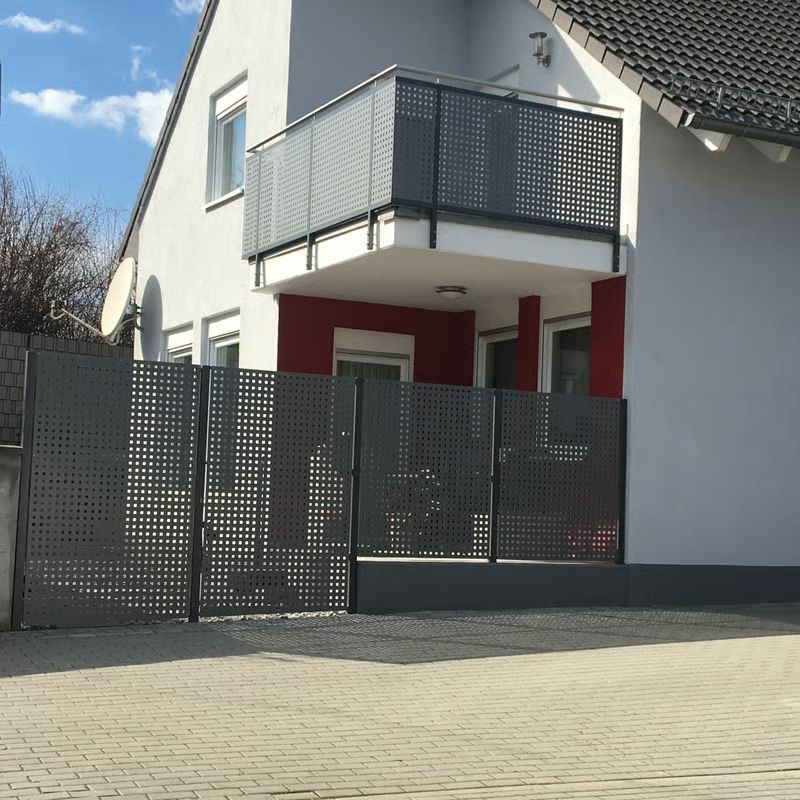 Modernes zweistöckiges Haus mit grauem Balkon, Metallzaun und gepflasterter Auffahrt unter blauem Himmel