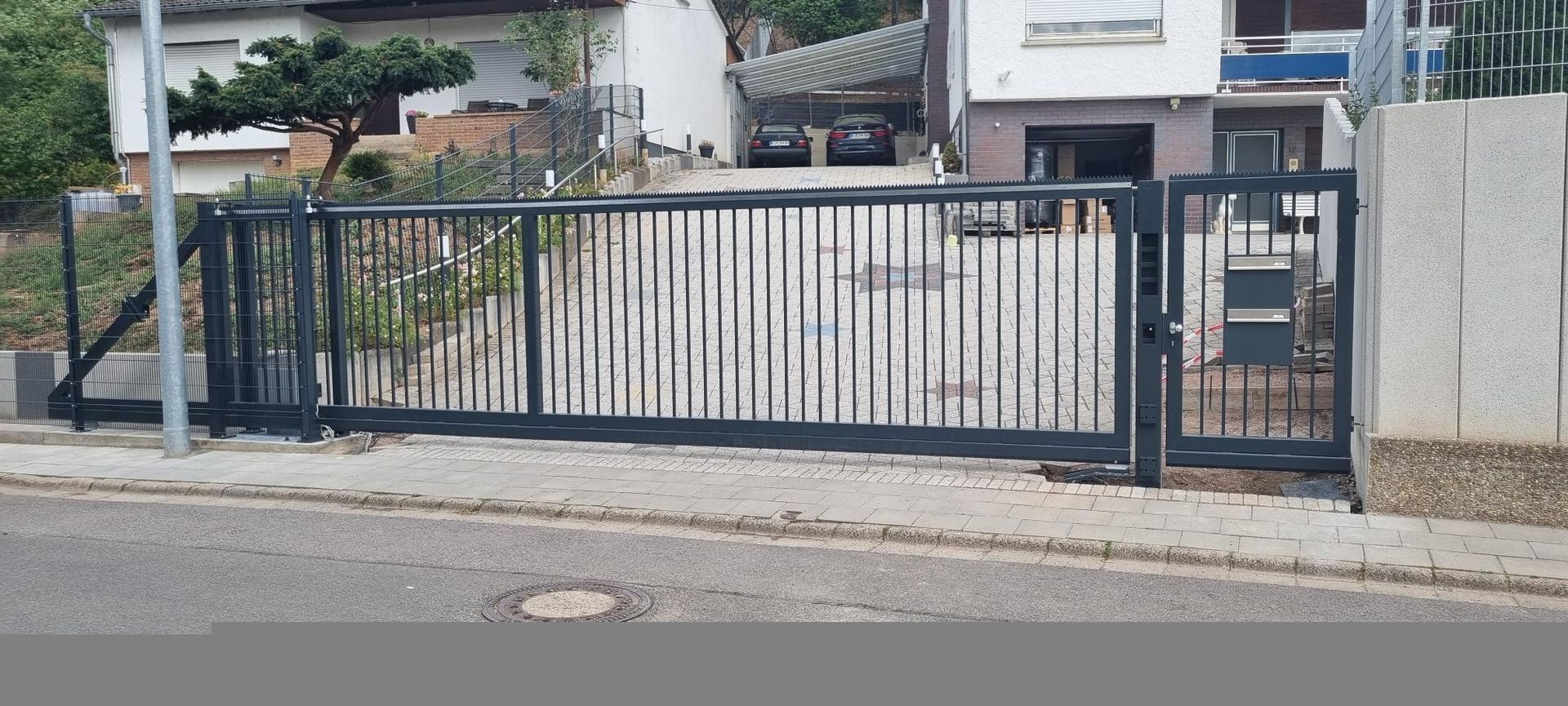 Schwarzes Metalltor und Zaun vor einer Wohnhauseinfahrt, dahinter Betonmauern und Gebäude.