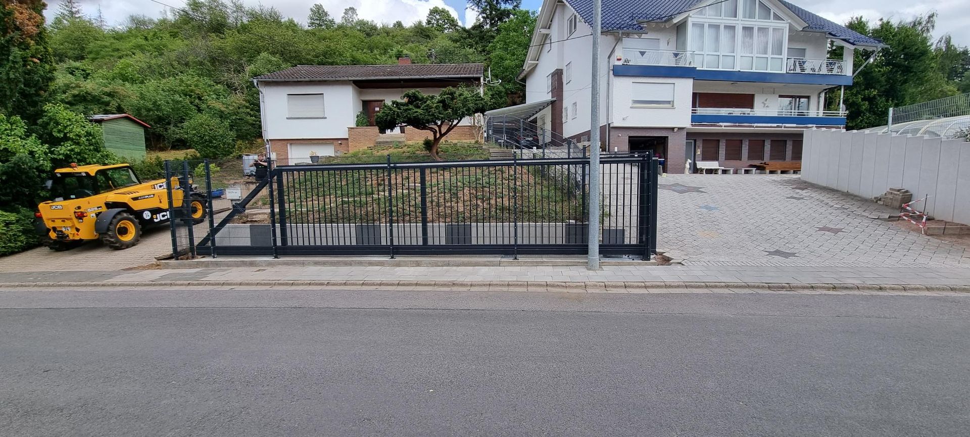 Wohnhaus mit schwarzem Metalltor und Kiesauffahrt, links daneben ein gelber Bagger geparkt.