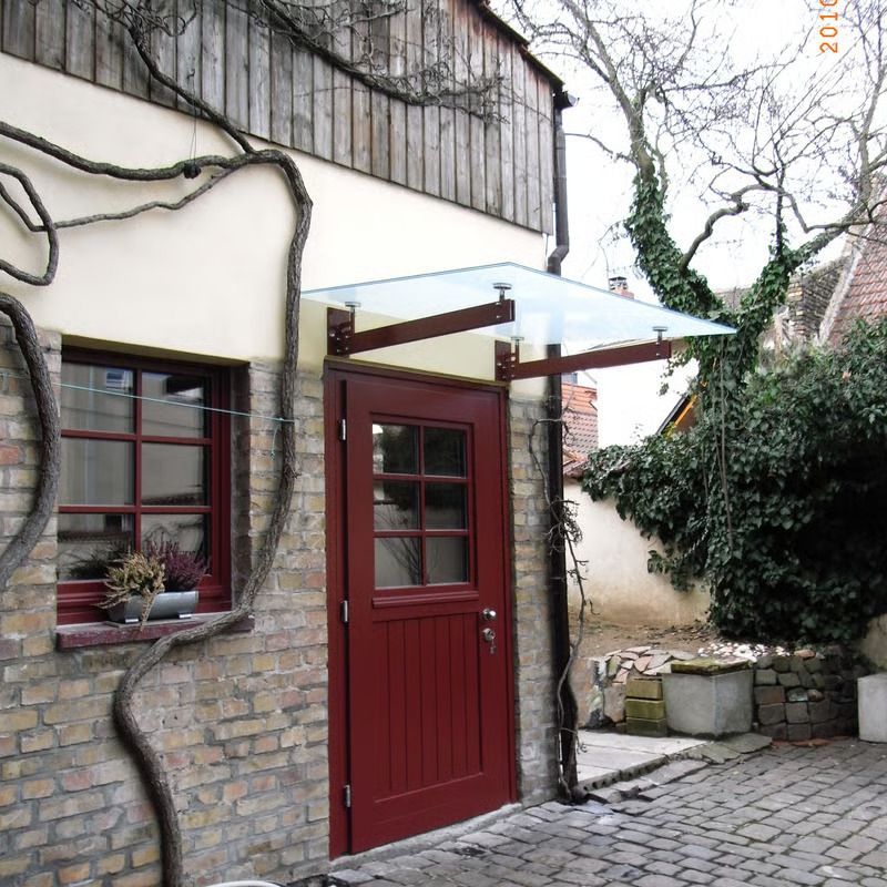 Rote Tür und Fenster an einem kleinen Steingebäude mit Glasvordach und Kopfsteinpflasterhof.