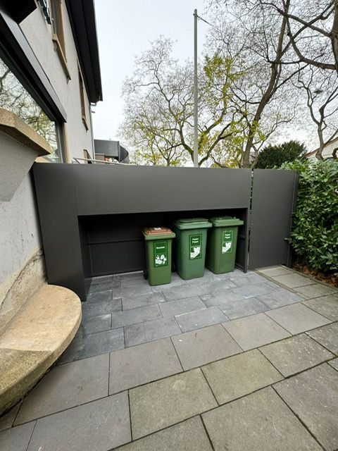 Drei grüne Mülltonnen neben einer grauen Mauer auf einer gepflasterten Außenterrasse