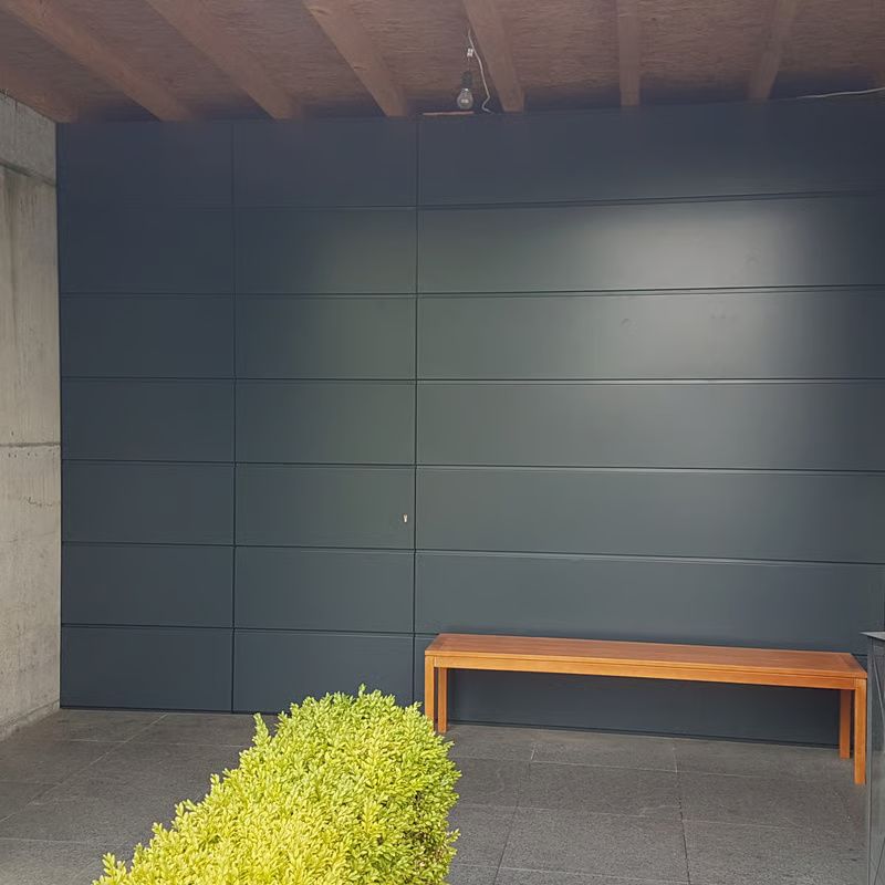 Moderne Terrasse mit dunkelgrauer Wand, Holzbank, Betonboden und einem grünen Strauch im Vordergrund