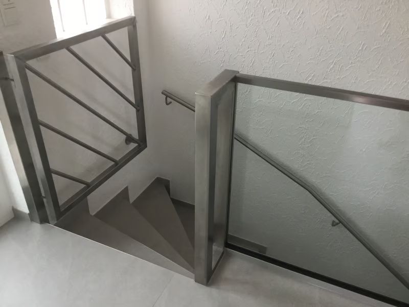 Treppenhaus mit grauem Metallgeländer und Glaspaneelen, das an einer weißen Wand und einem Fenster vorbeiführt.