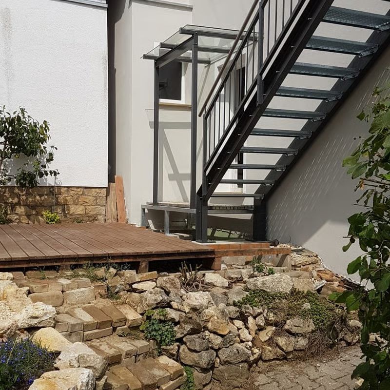 Außentreppe neben einem weißen Gebäude, im Vordergrund eine Holzterrasse und eine Steinstützmauer
