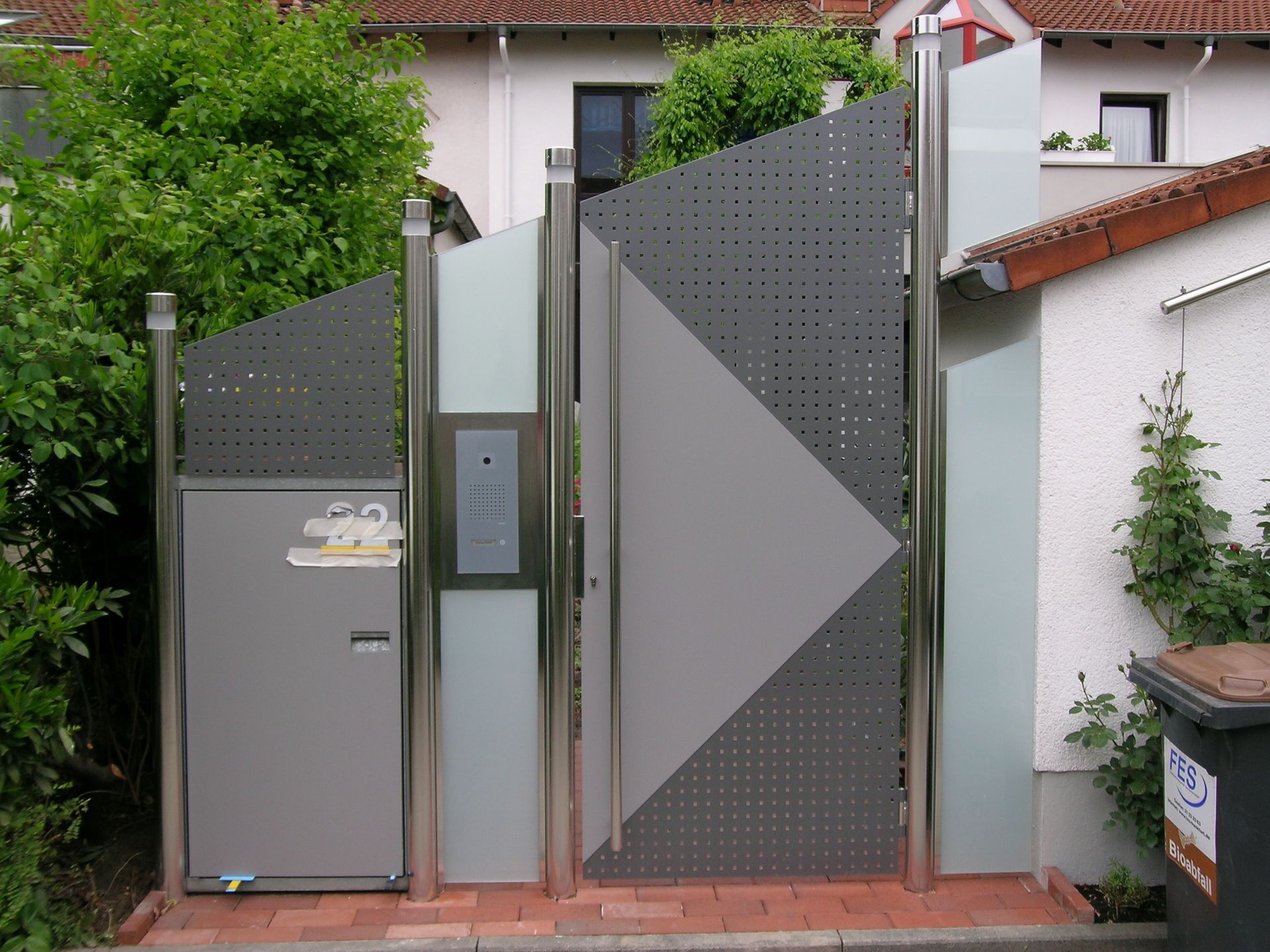 Modernes graues Metalltor und Briefkasten vor einem Haus mit grünen Hecken und rotem Ziegeldach