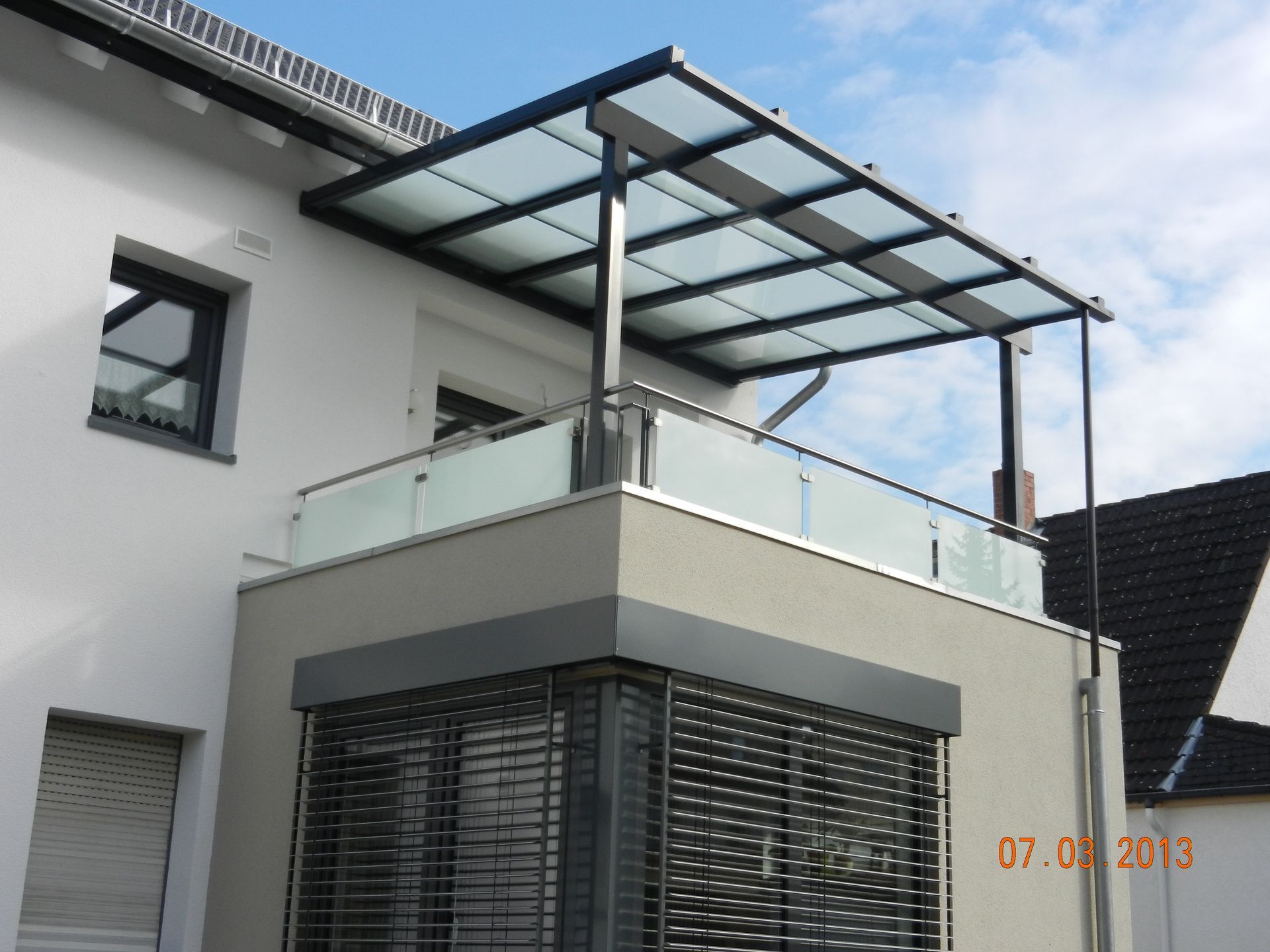 Modernes weißes Haus mit gläsernem Balkonvordach und dunklen Fensterläden unter einem teilweise bewölkten Himmel