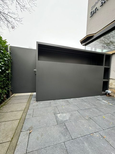 Graue, moderne Garage oder Schuppen mit Schrägdach neben einem gepflasterten Gehweg und an einer Gebäudeecke