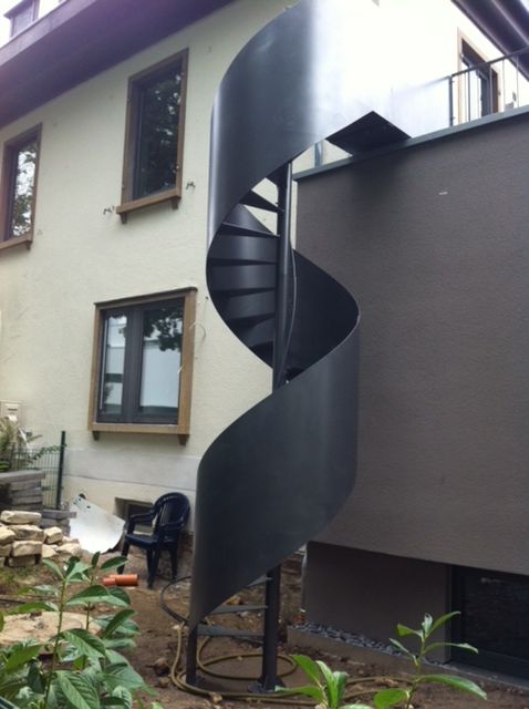 Schwarze, spiralförmige Außentreppe neben einem grauen Haus