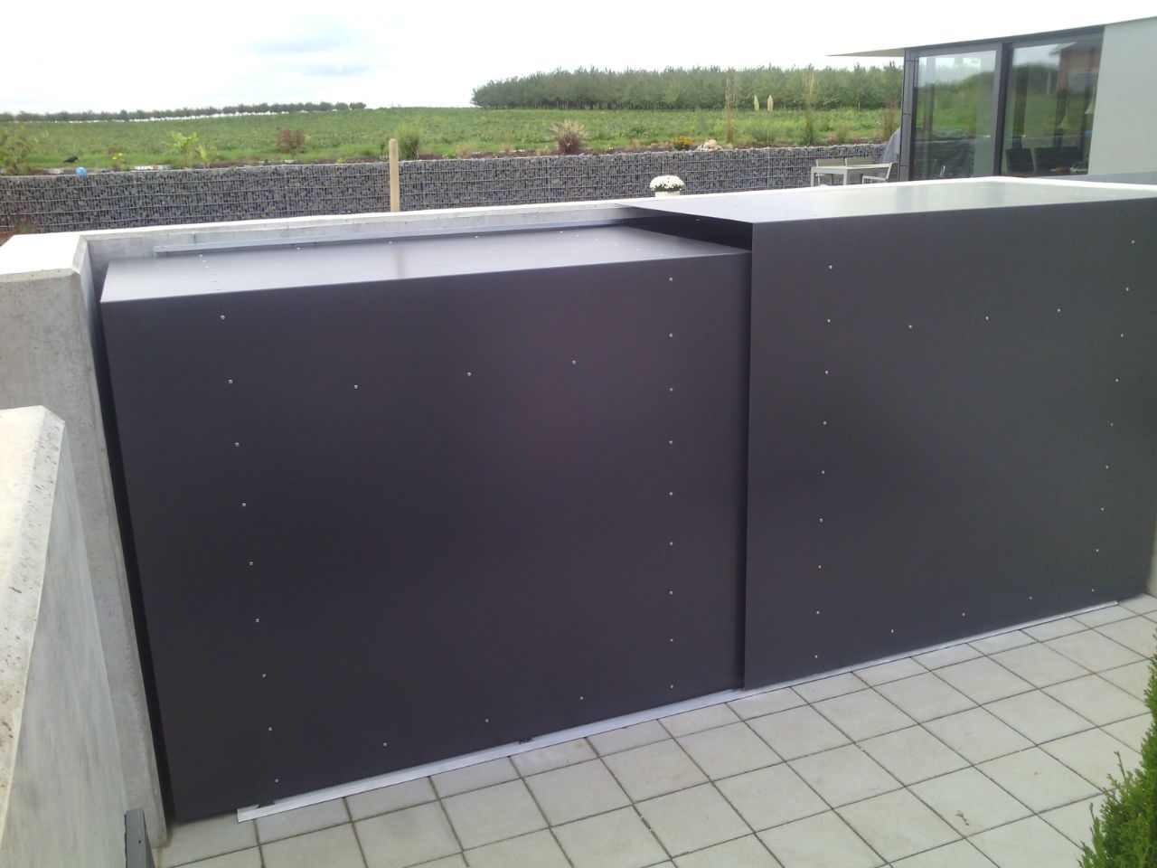 Schwarze Stromkästen neben einem gepflasterten Gehweg mit Blick auf ein grasbewachsenes Feld unter bewölktem Himmel
