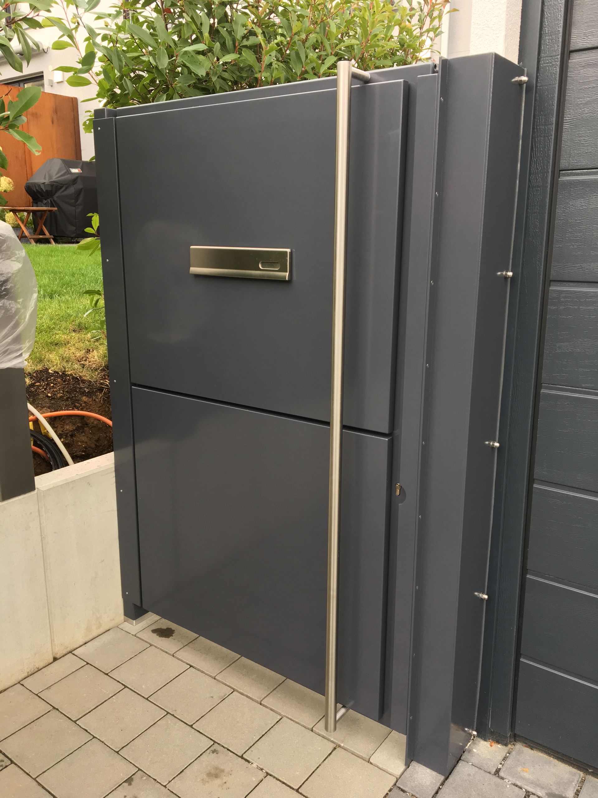 Dunkelgraues Eingangstor mit Briefkasten neben einem gepflasterten Gehweg und Gartenbegrünung