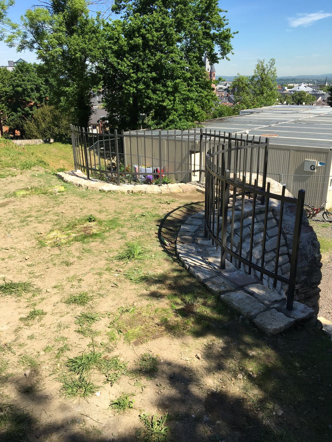 Hinterhof mit Baum, grasbewachsenem Hang und schwarzem Metallzaun neben einem kleinen Schuppen