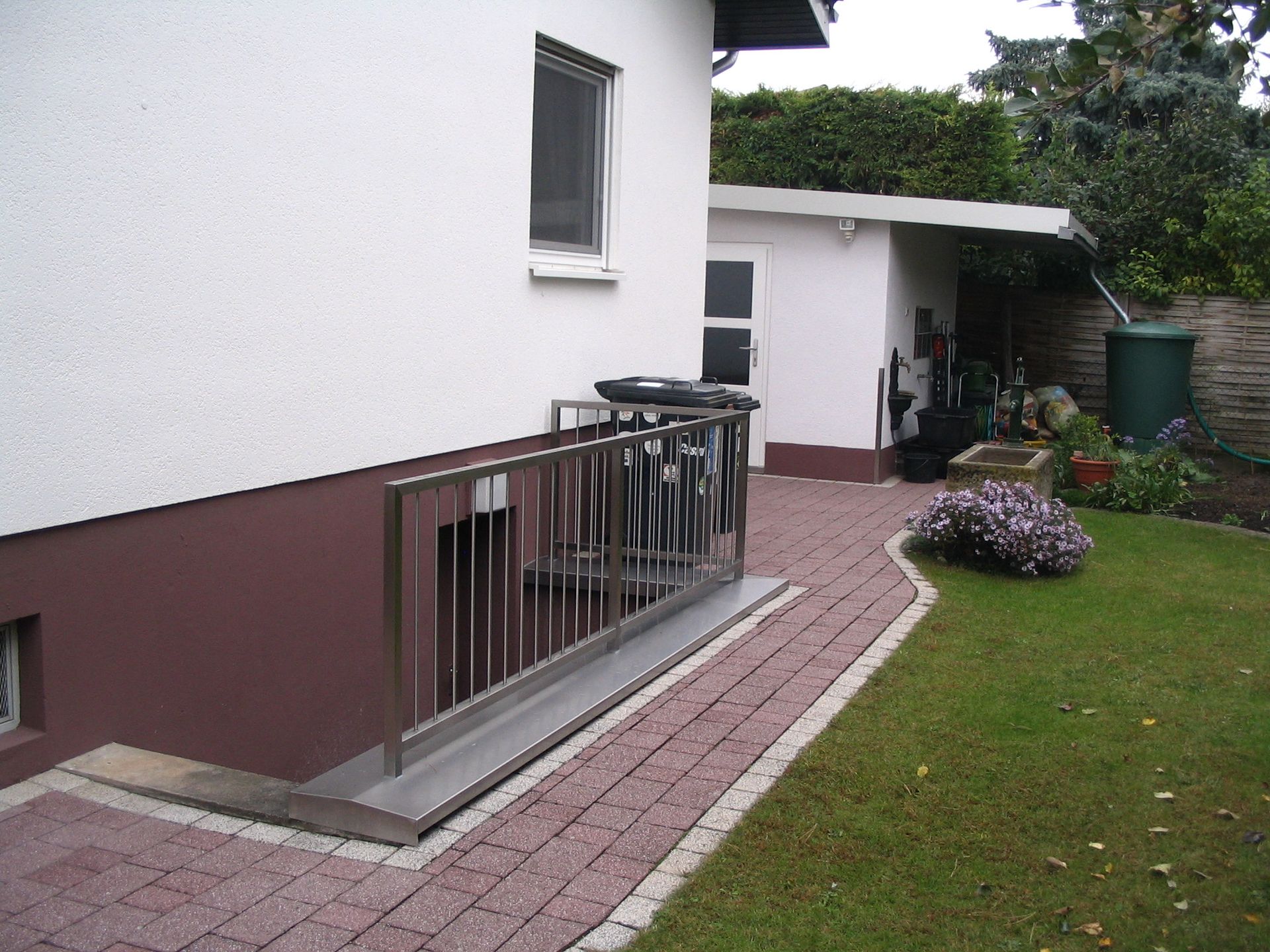 Außenansicht eines Hauses mit einer gemauerten Rampe, einem Metallgeländer und einem Rasen neben einer weißen Mauer