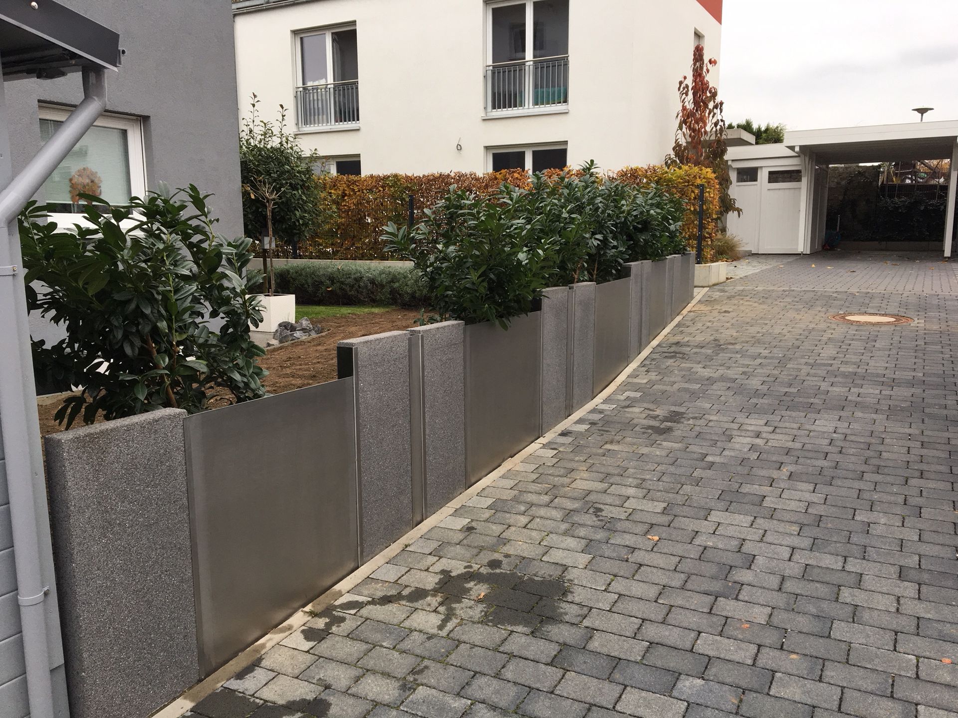 Kopfsteinpflasterauffahrt neben einem modernen Haus, umgeben von grauen Sichtschutzzäunen und Hecken.