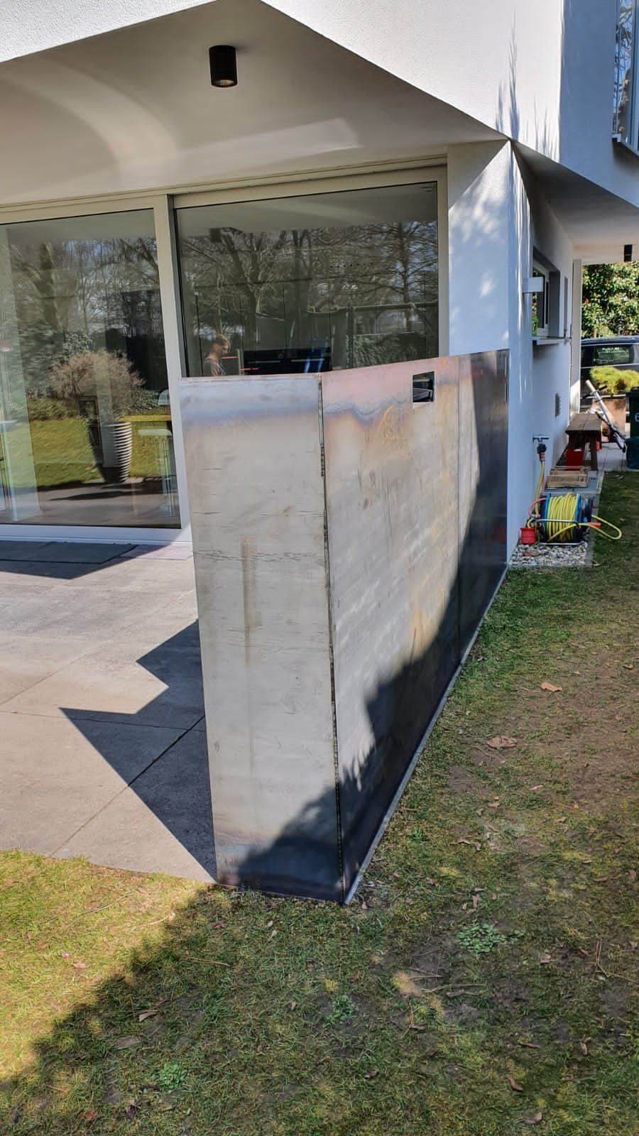 Betonstützmauer neben einem Haus mit Glaswänden, im Vordergrund Rasen und Terrasse.