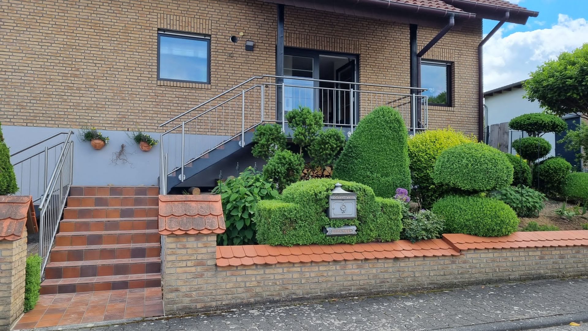 Vorderseite eines Backsteinhauses mit Treppe, gestutzten Sträuchern und einem kleinen Gartenschild am Gehweg