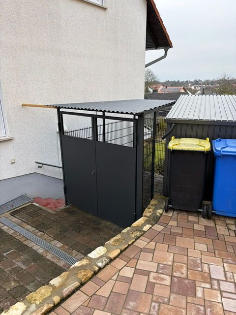 Abstellschuppen im Freien neben einem Haus, mit schwarzen Paneelen und einem Wellblechdach auf einer gepflasterten Terrasse.