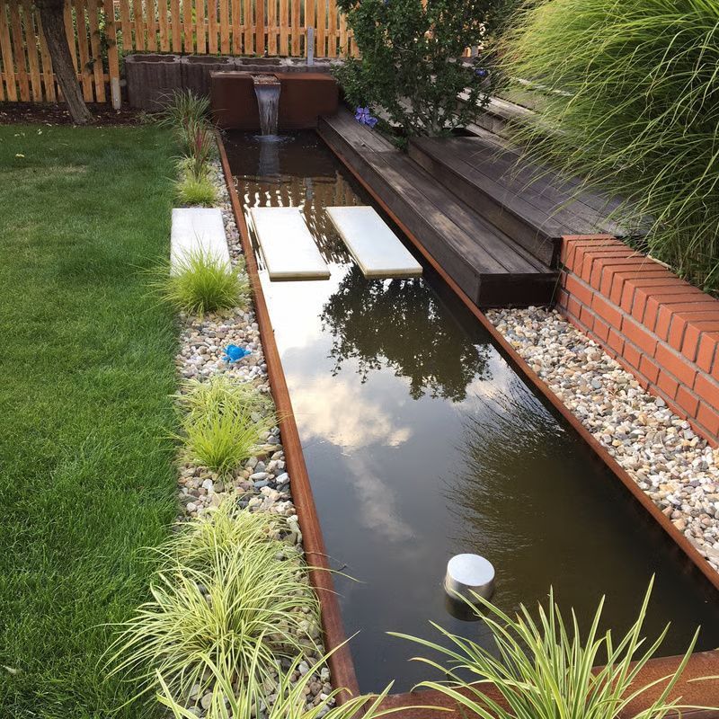 Schmaler, spiegelnder Gartenteich neben einer Holzterrasse und einer Backsteinmauer, mit Gräsern und kleinen Wasserfontänen.