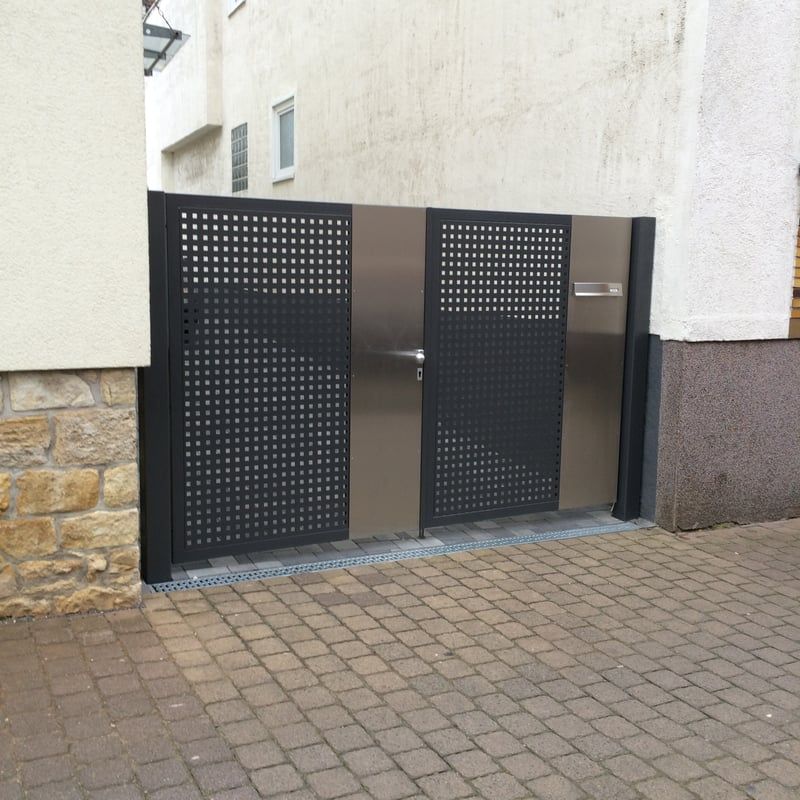 Schwarzes Metall-Fußgängertor in einem gepflasterten Eingangsbereich zwischen hellen Stuckwänden.