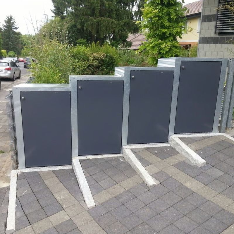 Vier dunkelgraue Mülltonnenkästen entlang eines Gehwegs neben geparkten Autos und Sträuchern.