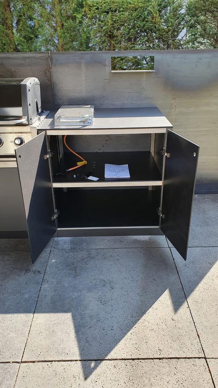 Offener schwarzer Schrank mit zwei Regalböden und weißer Arbeitsplatte auf einer sonnigen Terrasse.