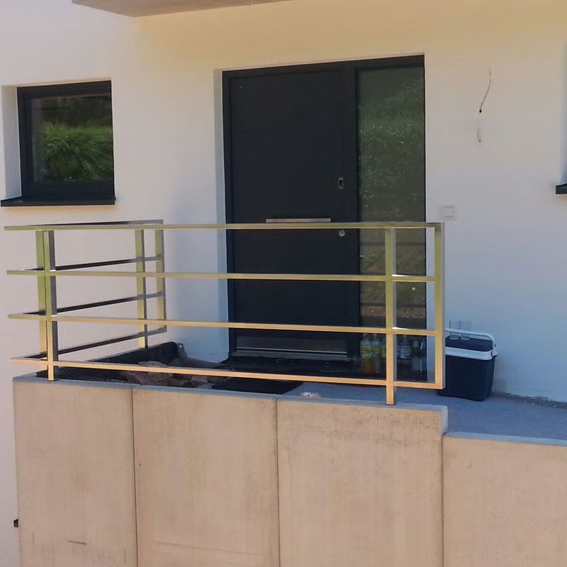 Außenbalkon mit goldenem Geländer, dunkler Tür und kleinem Fenster an einem weißen Gebäude.
