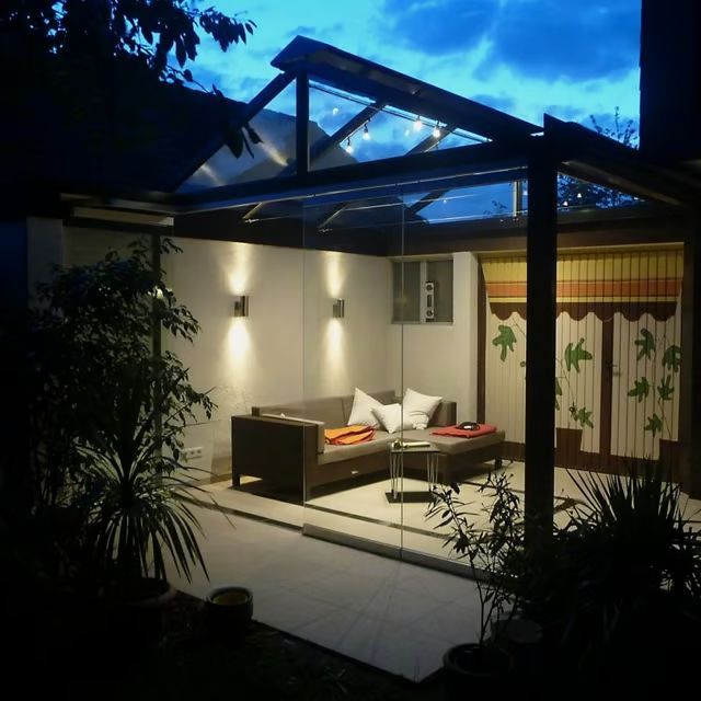Moderne Terrasse in der Abenddämmerung mit Glasdach, Sofa und stimmungsvoller Innen- und Außenbeleuchtung.