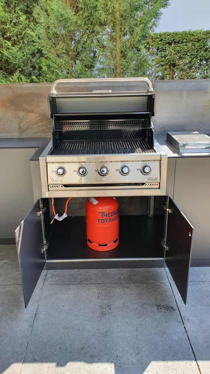 Gasgrill aus Edelstahl auf schwarzem Gestell im Freien mit einer roten Propangasflasche darunter.
