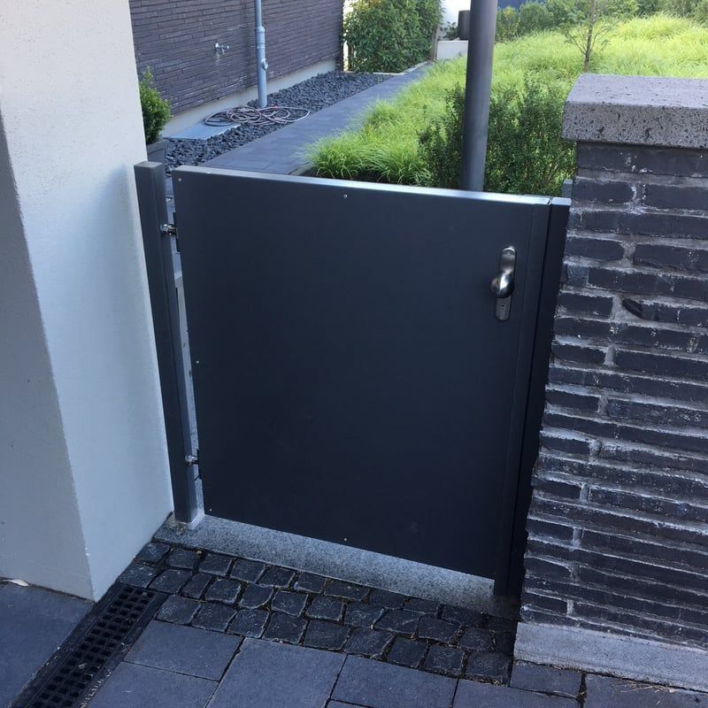 Ein kleines schwarzes Tor neben einer Backsteinmauer führt auf einen schmalen, gepflasterten Gartenweg.