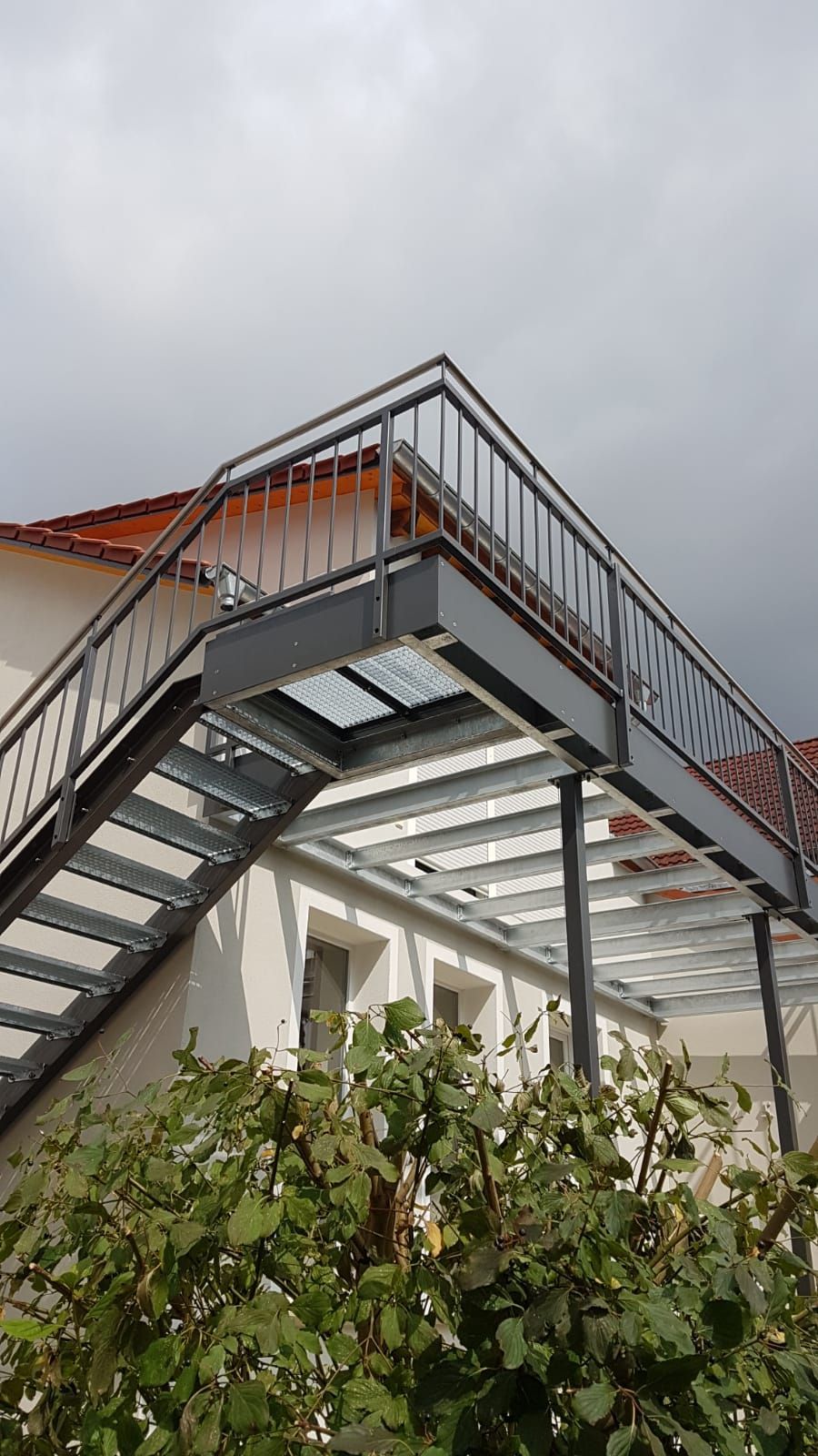 Außentreppe aus Metall und Balkon neben einem Gebäude, Blick über grüne Sträucher unter einem bewölkten Himmel