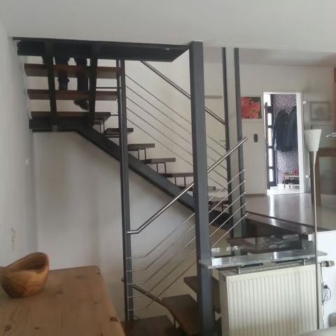 Moderne Innentreppe mit Metallgeländer in einem hellen Raum, neben einem Tisch und einem Heizkörper.