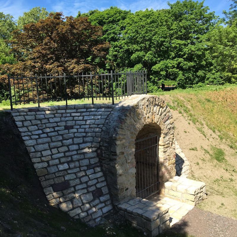 Steinbunker-Eingang mit Eisentor an einem grasbewachsenen Hang, unter Bäumen und strahlend blauem Himmel