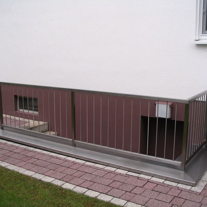 Kellereingang mit Metallgeländer entlang einer bordeauxroten Hauswand neben einem gepflasterten Gehweg und Rasen.